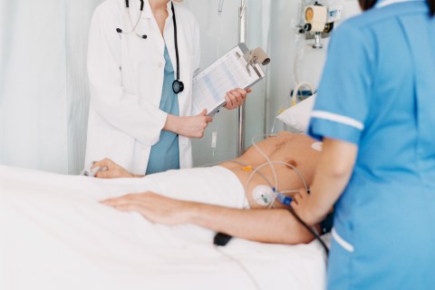 Female Doctor With a Clipboard and a Nurse Stand by a Male Patient on a Heart Rate Monitor